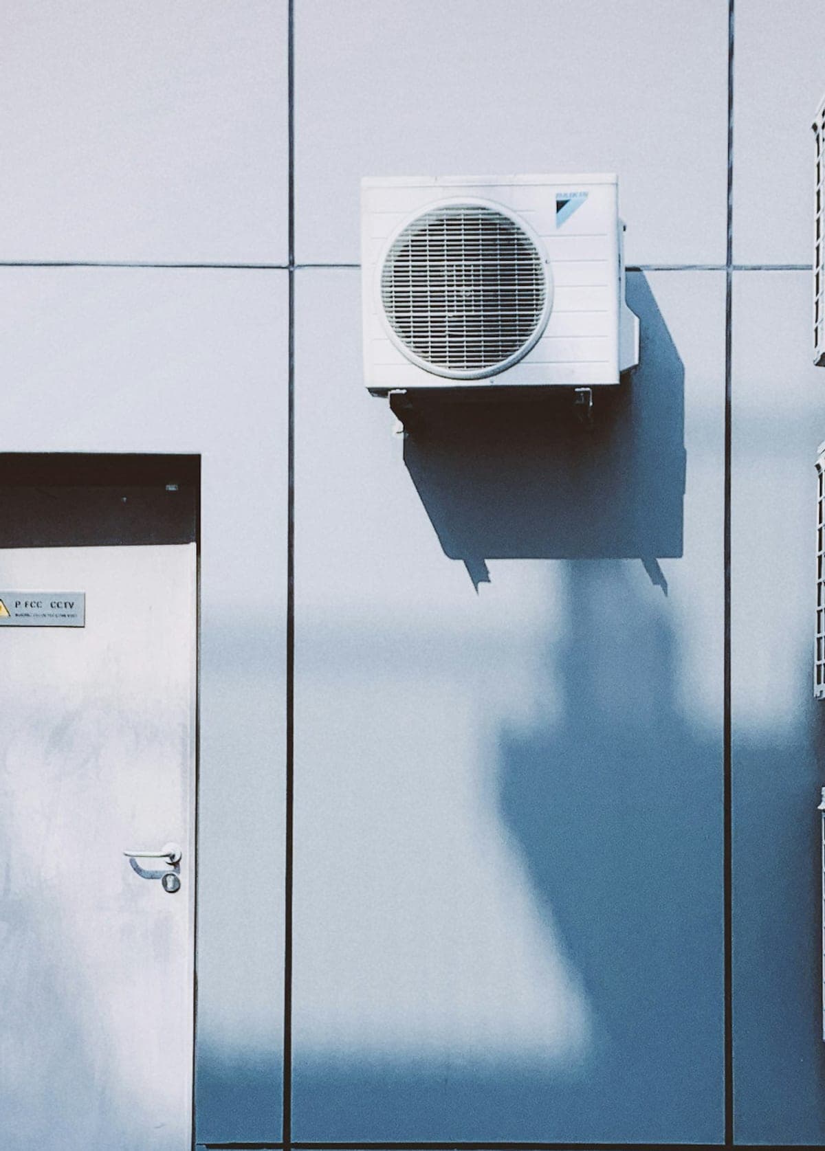 Unité extérieure de climatisation et sa grille de ventilation, fixée sur un mur à panneaux.
